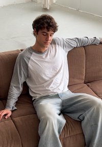 Hnědá látková pohovka s měkkou texturou; osoba v šedobílém tričku sedí uvolněně, má na sobě volné světle modré džíny.