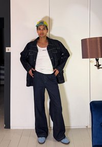 Νεαρό άτομο με μπούκλια, φοράει κίτρινο καπέλο, μαύρο τζιν μπουφάν, λευκό τοπ Versace, φαρδιά μπλε σκούρα παντελόνια και ανοιχτά μπλε παπούτσια μέσα σε κλειστό χώρο.