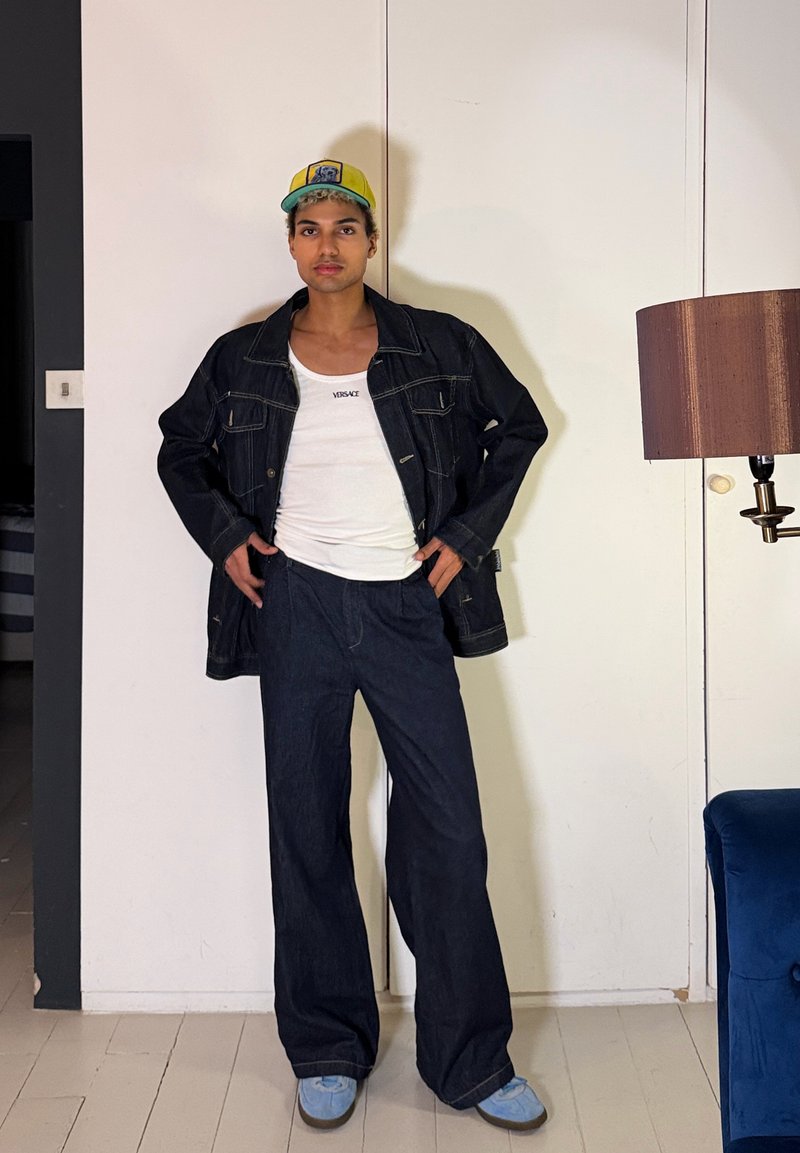 Jeune personne aux cheveux bouclés portant une casquette jaune, une veste en jean noire, un débardeur blanc Versace, un pantalon large bleu marine et des chaussures bleu clair à l'intérieur.