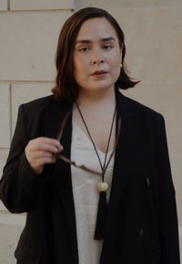 Blazer doppiopetto nero sopra una maglietta bianca. Una lunga collana con una perla rotonda e un tassello. La persona tiene degli occhiali da sole in una mano.