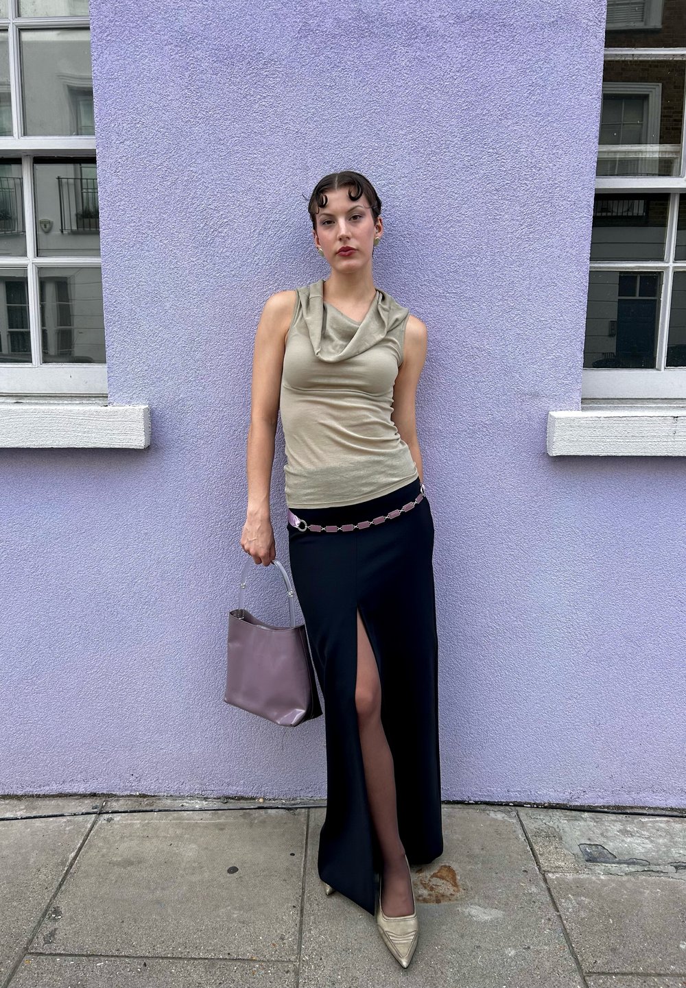 Μεταλλική μπλούζα με γιακά, μαύρη φούστα με πλαϊνό σκίσιμο, ζεμπίλι με ροζ ζώνη, κρατώντας μία καφέ τσάντα, καφέ μυτερές γόβες, μπροστά σε έναν μωβ τοίχο.