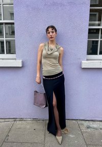 Μεταλλική μπλούζα με γιακά, μαύρη φούστα με πλαϊνό σκίσιμο, ζεμπίλι με ροζ ζώνη, κρατώντας μία καφέ τσάντα, καφέ μυτερές γόβες, μπροστά σε έναν μωβ τοίχο.