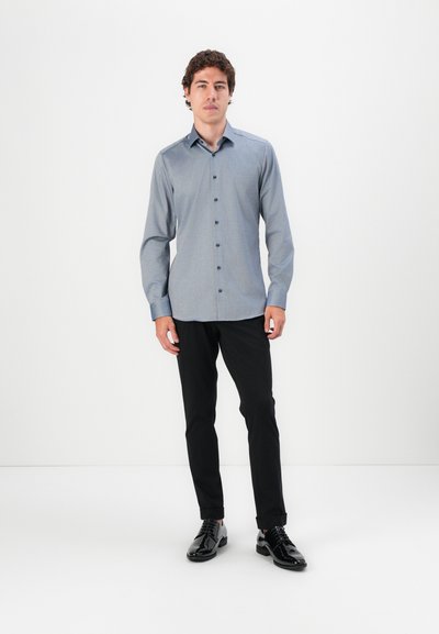 Camisa azul claro de botones con una textura sutil, combinada con pantalones negros ajustados y zapatos de vestir brillantes en negro.