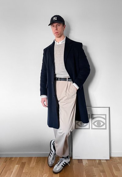 Hombre con abrigo negro, suéter gris, pantalones beige, zapatillas deportivas y gorra negra apoyado contra una pared blanca cerca de una obra de arte enmarcada con un ojo.