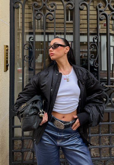 Chaqueta bomber negra sobre un top corto blanco, combinada con vaqueros anchos de pierna brillante en color azul y un bolso negro con detalles de herrajes plateados.