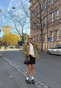 Olivo zelena jakna čez belo jopico, črna mini krilo, bele nogavice in črne platformne čevlje. Rjava torba, stoji na ulici s drevesi.