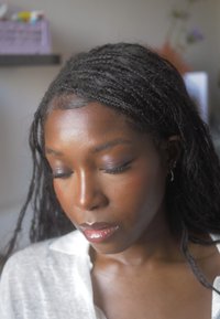 Retrato de una persona con cabello negro largo y trenzado, que lleva una camisa blanca ligeramente arrugada. Presenta un tono de piel natural y labios brillantes.