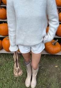 Osoba u bijelom puloveru, bijelim šortsevima i bež čizmama stoji na travi držeći bež torbicu nemo blizu n stacked bundeva na drvenim policama.