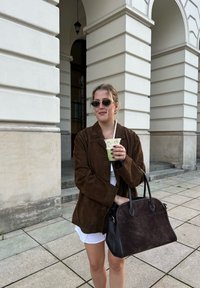 Barna szarvasbőr kabát, fehér ing, átlátszó pohárban zöld italt tartva. Fekete táska arany díszítéssel. Négyzet alakú csempék és kőoszlopok a háttérben.