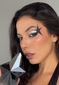 Svart stjärnformat parfymflaska med en blank yta hålls nära ansiktet som är sminkat med silver och svart ögonskugga.