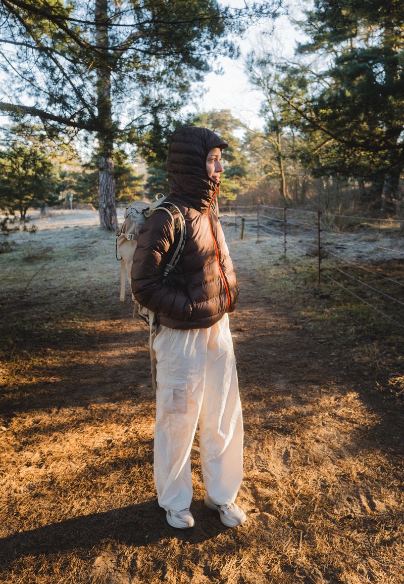 Doudoune marron avec capuche, fermeture éclair orange et sac à dos kaki. Pantalon cargo blanc ample et chaussures claires. Cadre naturel en plein air.