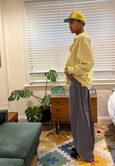 Camisa de cuadros amarillos oversize, pantalones grises de pierna ancha, zapatos negros con rayas amarillas, gorra verde, entorno interior con una planta y una alfombra colorida.