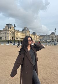 Γυναίκα με μακριά σκούρα μαλλιά και γυαλιά που φορά καφέ παλτό και γκρι πουλόβερ, στέκεται κοντά στο Μουσείο του Λούβρου και την γυάλινη πυραμίδα κάτω από συννεφιασμένο ουρανό.