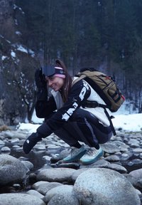 Une personne est accroupie sur des pierres lisses près d'un ruisseau, portant une veste noire et ivoire, des gants noirs et des chaussures de sport bleu clair.