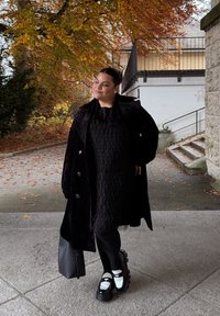 Černý, texturovaný dlouhý kabát přes vrstvené černé šaty s třásněmi. Černé punčocháče a masivní černo-bílé platformové boty. Černá taška.