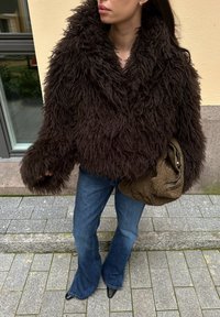 Hnedá kožušinová bunda s chumáčmi, voľný oversized strih, s hlbokým výstrihom do V. Kombinovaná s modrými zvonovými džínsami a texturovanou hnedou kabelkou.