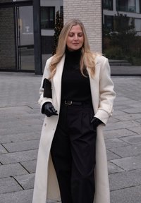 Biely vlnený kabát cez čierny rolák, doplnený čiernymi nohavicami. Čierne kožené rukavice a čierna kabelka dopĺňajú outfit.