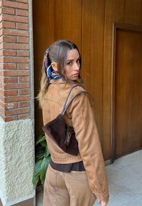 Brązowa zamszowa kurtka z ćwiekami, noszona na brązowych spodniach sztruksowych. Na ramieniu zawieszona ciemna torba, we włosach kolorowa chusta.