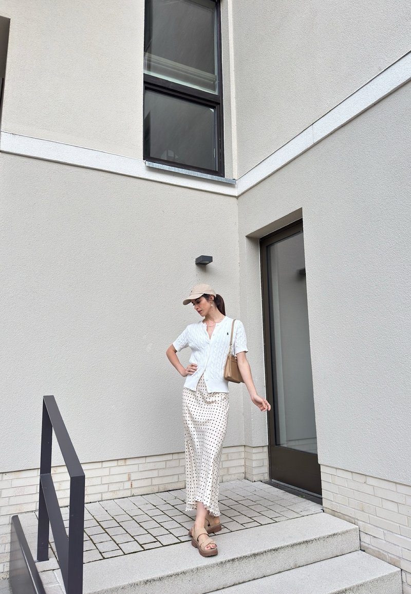 Woman wearing a white cardigan, polka dot skirt, beige cap, and sandals standing on steps outside a modern building near a glass door.