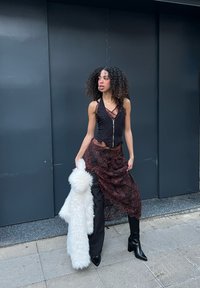 Musta vartalonmyötäinen paita vetoketjulla, kerroksittainen pitsihame tummanpunaisena, mustat housut ja polveen ulottuvat mustat saappaat, pitää kädessään pörröistä valkoista hartiahuivia.