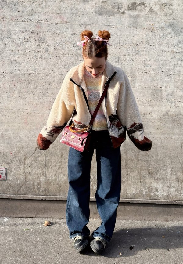 Veste en polaire avec un corps crème et des accents marron, pull graphique, jean large bleu, sac à main rose à motif, chaussures foncées.