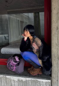 Jeune femme en collants bleu vif, chaussures à imprimé léopard et manteau en fourrure accroupie près d'un mur en béton, avec un sac Hello Kitty violet à côté d'elle.
