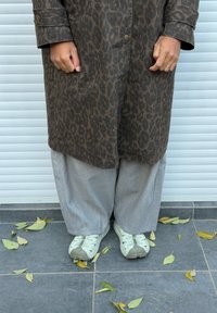 Manteau imprimé léopard marron avec de gros boutons, pantalon gris à jambes larges, et baskets vert clair avec un design texturé, posés sur un sol carrelé avec des feuilles.