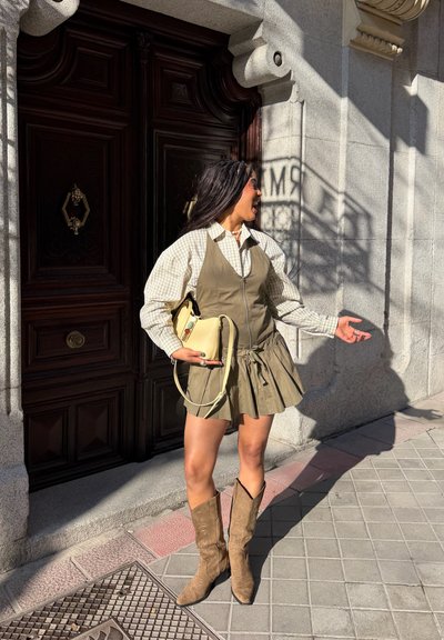 Joven mujer con vestido beige, camisa de cuadros y botas, de pie en la acera sosteniendo un bolso crema, gesticulando y sonriendo hacia la derecha.