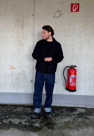 Chaqueta negra sobre una camisa negra, jeans azules con un puño azul claro, zapatos negros. Extintor de incendios rojo montado en la pared gris.