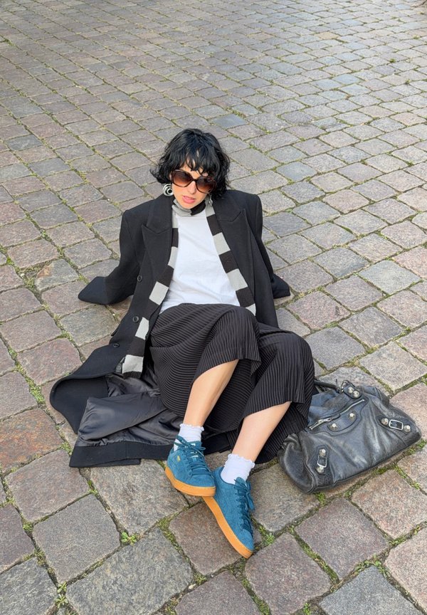 Mujer con cabello rizado negro y gafas de sol sentada sobre un suelo de adoquines, vestida con un abrigo negro, bufanda a rayas, falda plisada y zapatillas azules.