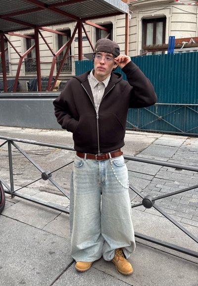 Joven con vaqueros claros de gran tamaño, sudadera marrón con cremallera, camisa de cuadros, botas color canela y gorra puesta de lado, de pie junto a una barandilla metálica en una acera urbana.