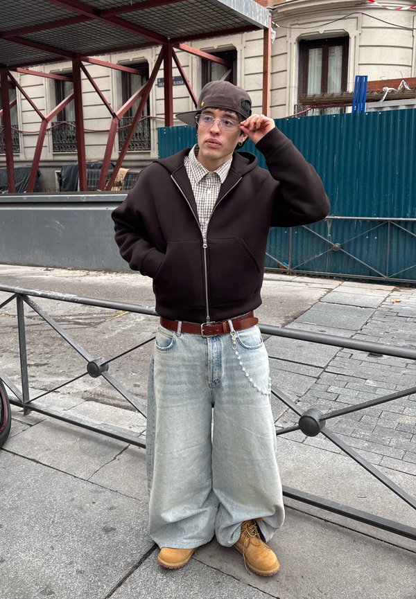 Young person in oversized light jeans, brown zip-up hoodie, checked shirt, tan boots, and sideways cap stands near metal railing on urban pavement.