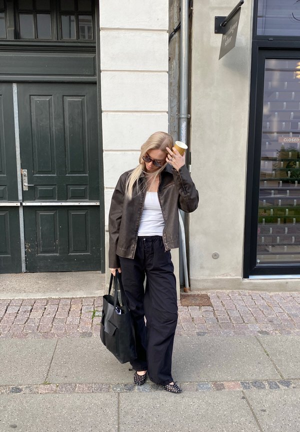Chaqueta de cuero marrón, top blanco sin mangas, pantalones ancho negros, bolso negro, zapatos de lunares, gafas de sol, sosteniendo una taza, entorno urbano.