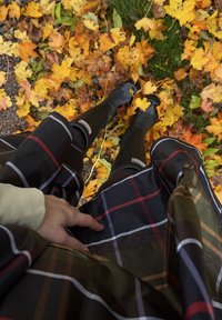 Un impermeabile a quadri neri e multicolori pende su stivali di gomma neri, sistemato su un terreno di foglie autunnali gialle e arancioni.