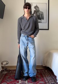 Γκρι πλεκτό πουλόβερ, ανοιχτό μπλε φαρδιά τζιν, μαύρες ξυλοπόδαρες, κρατώντας μια μαύρη δερμάτινη τσάντα. Ο φόντος περιλαμβάνει μια κορνιζαρισμένη μονοχρωματική αφίσα και μια λάμπα.