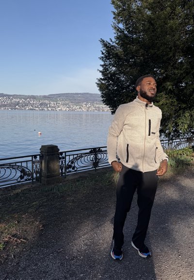 Hombre con chaqueta clara y zapatillas de deporte de pie en un camino junto al lago cerca de una barandilla, con agua y colinas lejanas bajo un cielo despejado.