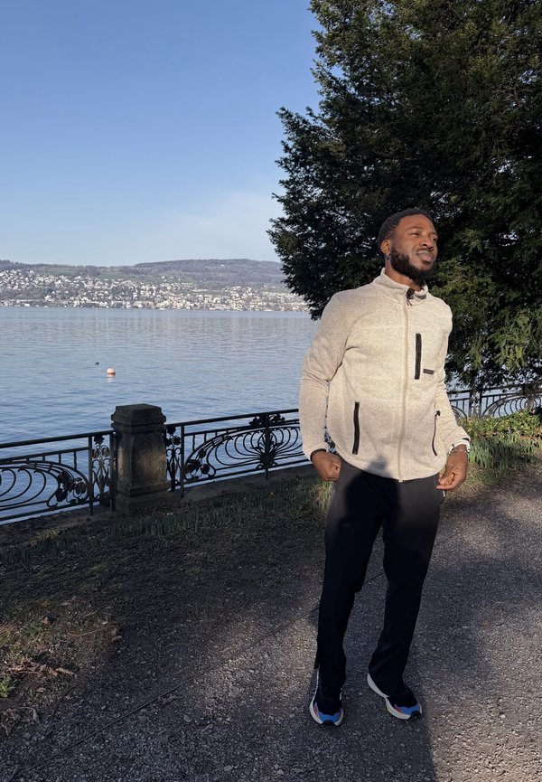 Homme en veste claire et baskets debout sur un sentier au bord du lac près d'une rambarde, avec de l'eau et des collines lointaines sous un ciel clair.