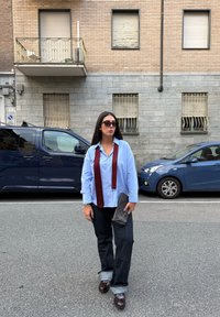 Ανοιχτό μπλε πουκάμισο με γιακά και μπορντό γραβάτα, σκούρο τζιν με σηκωμένα μπατζάκια και καφέ παπούτσια. Κρατάει μια σκούρα τυπωμένη τσάντα. 