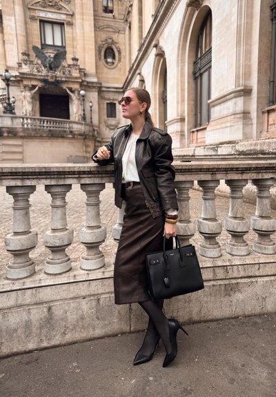 Mujer con gafas de sol, chaqueta de cuero negra, blusa blanca, falda oscura y tacones, apoyada en una balaustrada de piedra cerca de un edificio histórico.