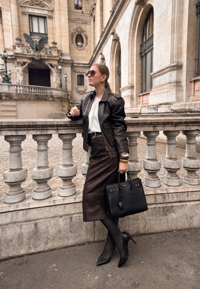 Mujer con gafas de sol, chaqueta de cuero negra, blusa blanca, falda oscura y tacones, apoyada en una balaustrada de piedra cerca de un edificio histórico.