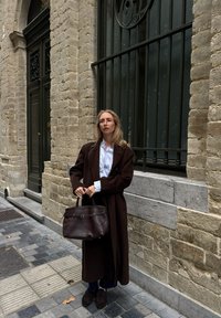 Cappotto di lana marrone sopra una camicia bianca, tiene una borsa di pelle marrone scuro. Muro di pietra e barre alle finestre in un contesto urbano, con pavimento in ciottoli.