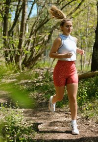 Abbigliamento sportivo composto da una maglietta senza maniche azzurro chiaro e pantaloni corti rossi a fantasia. Scarpe da corsa bianche con accenti grigi, che corrono su un sentiero di terra.