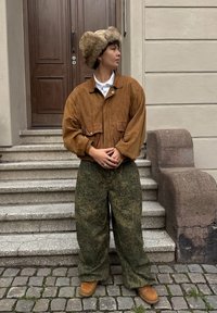 Brązowa zamszowa kurtka, kremowy futrzany kapelusz z klapkami na uszy, zielone spodnie w wzory i brązowe buty, stojący na kamiennych schodach.