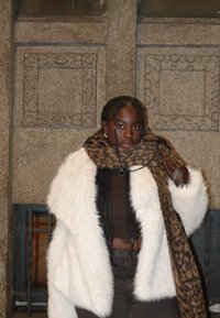 Femme aux cheveux tressés portant un manteau en fourrure blanche, un haut marron, une écharpe léopard, un gant en cuir, debout contre un mur en pierre orné la nuit.