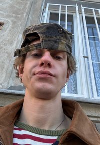 Kasket med camouflage, bøjet fremad, stribet sweater med grøn kant, brun frakke og øreringe, mod en baggrund af et vindue.