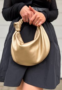 Borsa in finta pelle dorata con manico annodato, texture morbida e forma arrotondata, tenuta da una persona in una gonna nera.