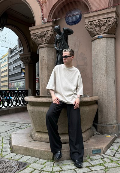 Camisa de algodón beige de gran tamaño, pantalones anchos negros y zapatos negros. Sentado cerca de una fuente de piedra y una estatua en un entorno urbano.