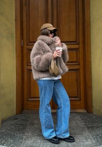 Műszőrme barna dzseki, világoskék farmerek, fekete platform cipők, bézs kézitáska, fehér kávéscsésze a kézben, sötét sapka és napszemüveg.