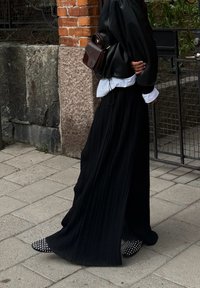 Czarna plisowana spódnica maxi, oversize'owa czarna bluzka, widoczne białe mankiety koszuli, czarne buty z bia�łymi ćwiekami, mała brązowa torba na ramię, kamienne tło.