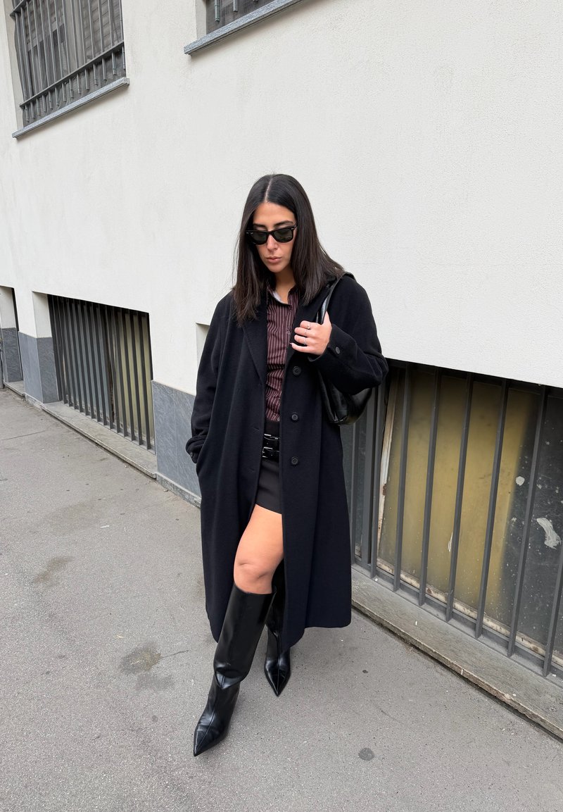 Casaco longo preto de lã, camisa castanha às riscas, minissaia preta, botas pretas pontiagudas até ao joelho e uma pequena mala preta. Ambiente urbano.